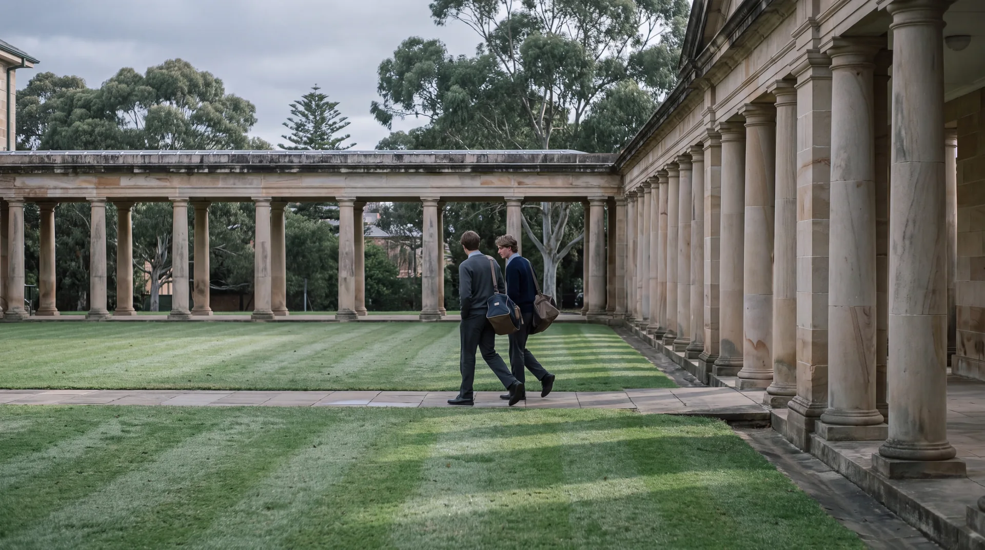 Two students walking through a sandstone school colonnade in early morning light.