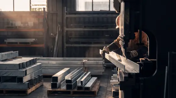 Gloved hands guiding a galvanised steel component off a press brake in a fabrication plant, light falling from high windows.