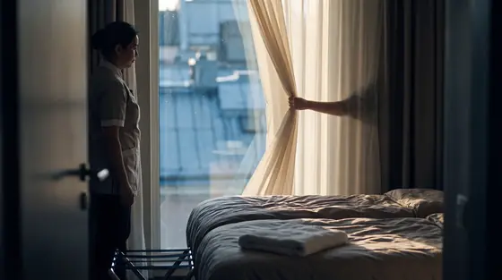 A housekeeper opening the curtain of a hotel room at dawn, light falling across a made bed.