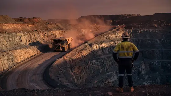 An operator in hi-vis watching a haul truck climb an open cut mine ramp at last light, red dust hanging in the air.