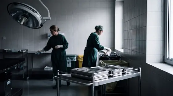Two theatre staff in green gowns turning over an operating theatre between cases, instrument trolley in the foreground under cold light.