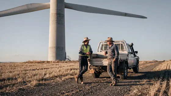 A project engagement officer and a farmer talking beside a ute on dry farmland below the base of a wind turbine in late afternoon light